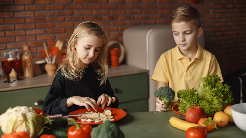 Positive little girl and boy sitting at desk with various fresh vegetables and fruits while preparing meal in loft kitchen with brick wall